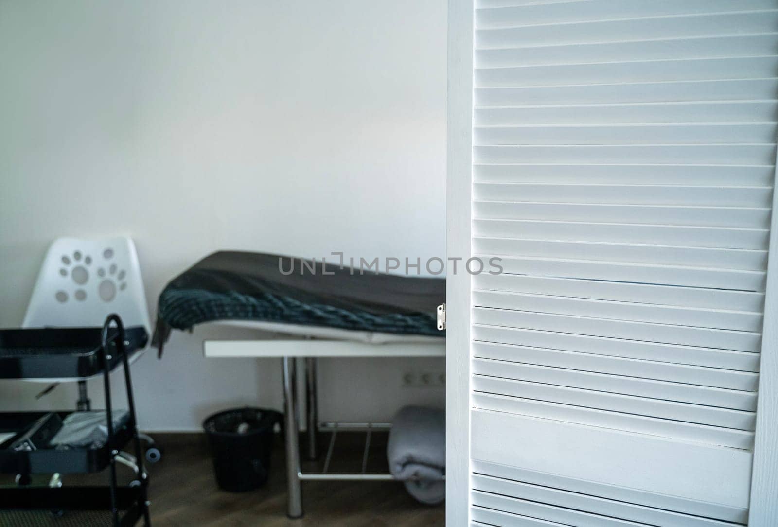table for massage and other cosmetology services. covered with a blanket and a disposable sheet. next to it is a white chair. in the foreground is a white screen.