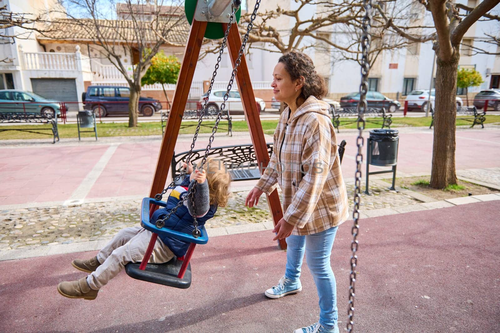A mother gently pushes her child on a swing at a playground on a sunny day. The outdoor park setting features play equipment, surrounding trees, and a cheerful scene of togetherness and connection.