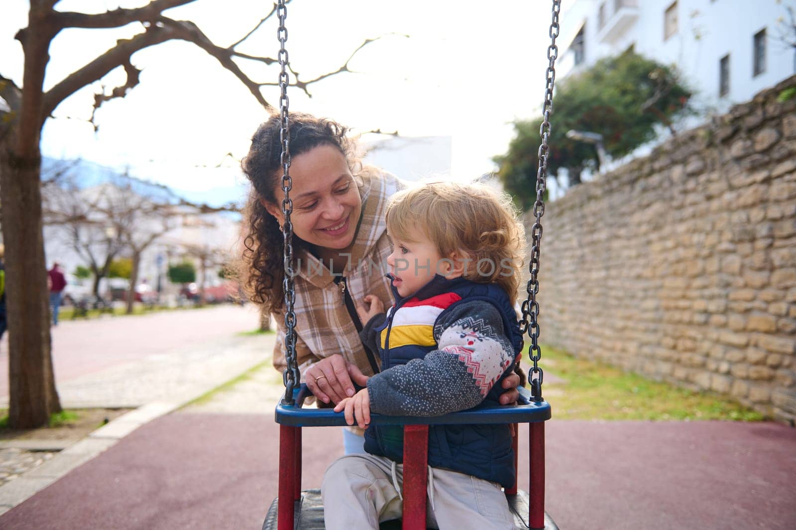 Happy moment of a mother engaging with her toddler on a swing in a park setting. The scene conveys warmth, family connection, and the joy of a shared outdoor activity.