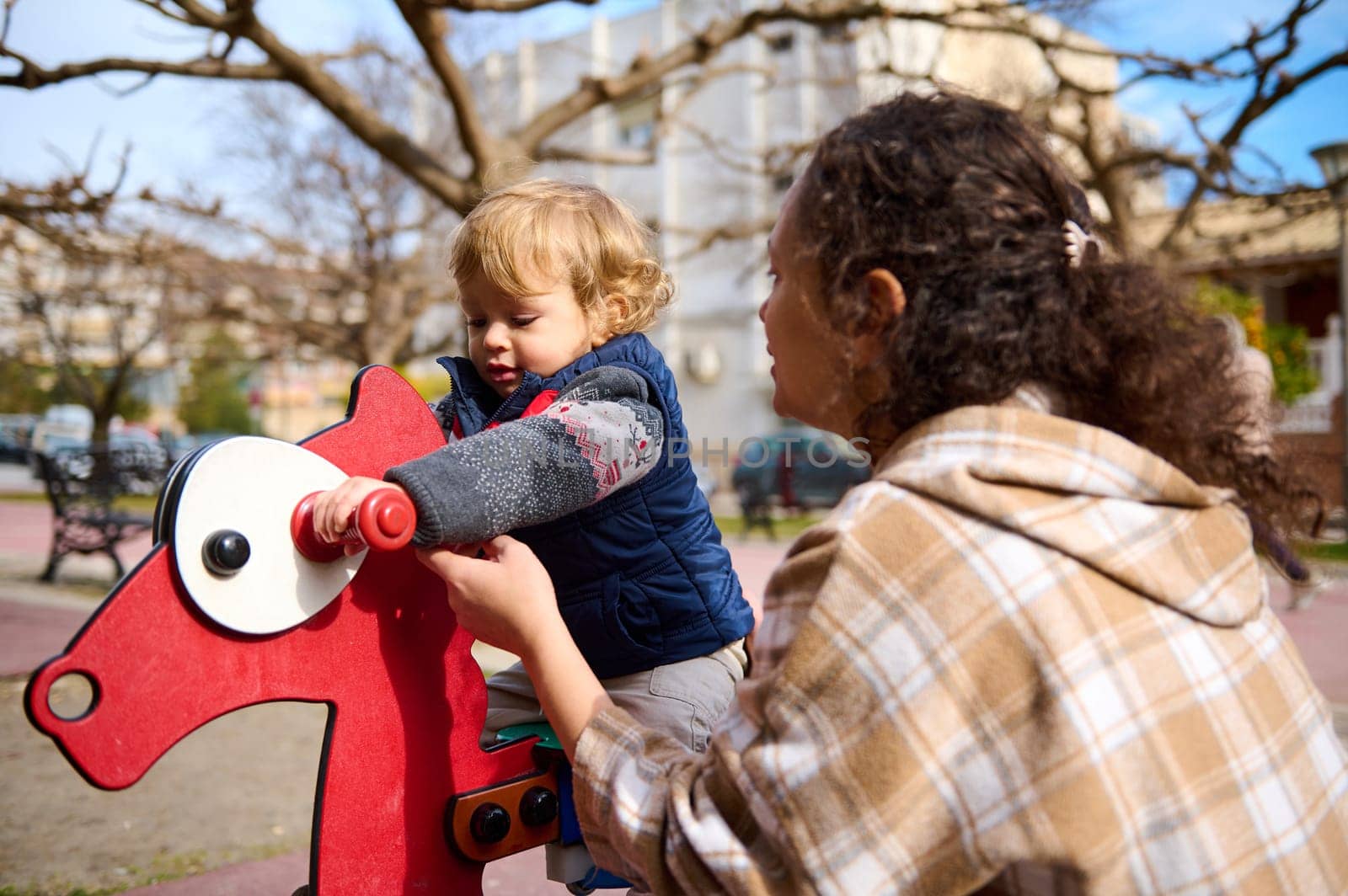 A caring mother is helping her young child ride a red rocking toy at a park. The scene depicts parenting, outdoor fun, and a moment of joy under sunny skies.