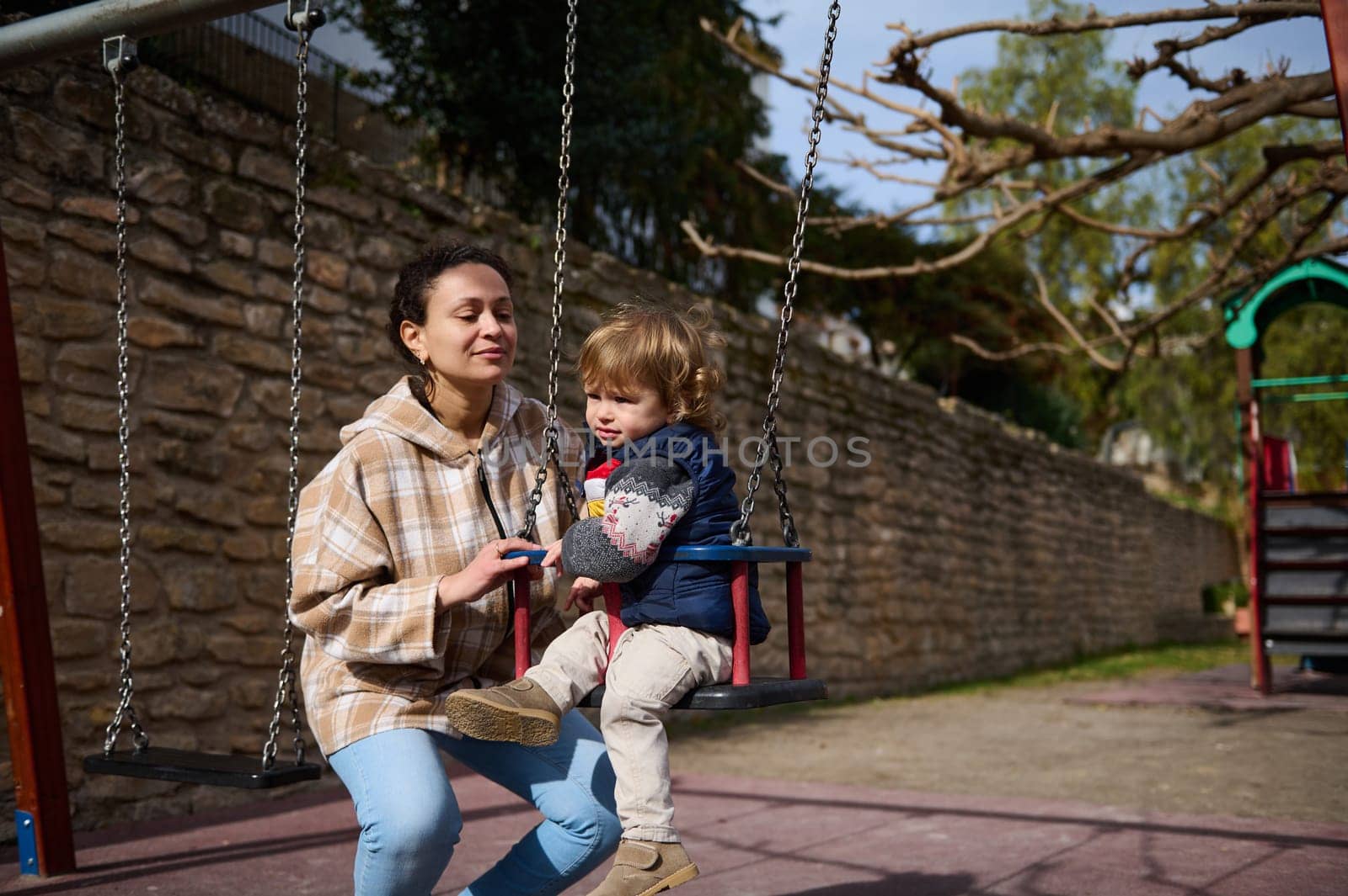 A mother and her young son bonding together in a park while using a swing on a sunny day.
