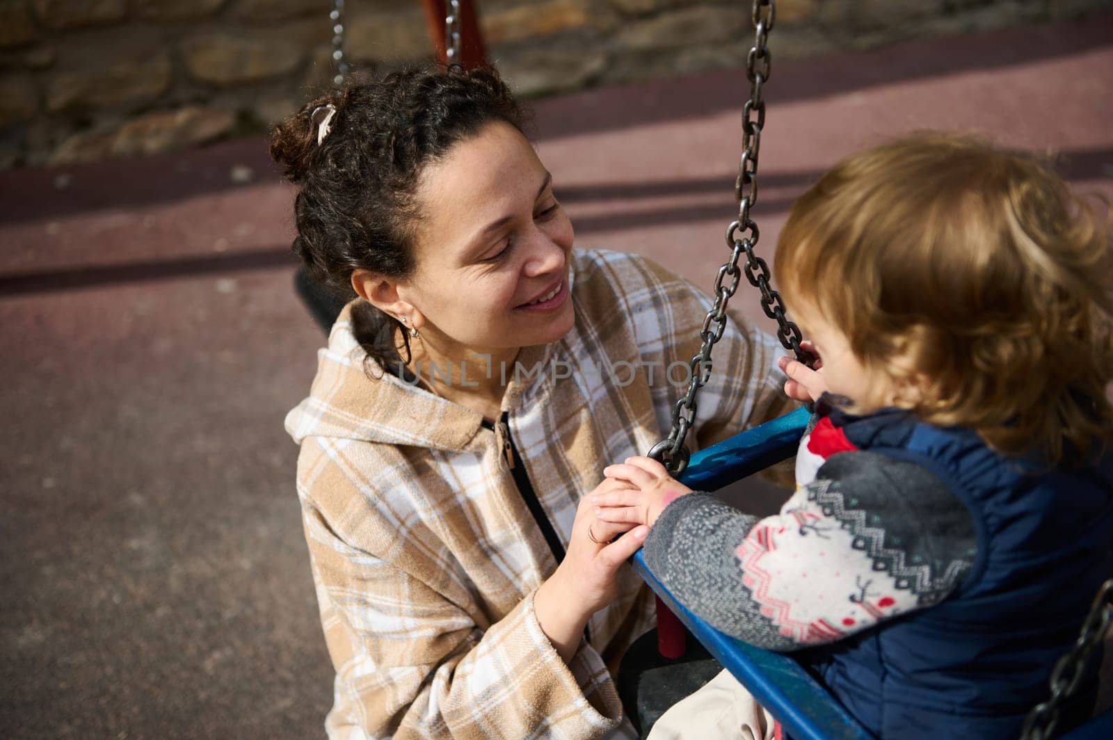 A mother lovingly interacts with her young child who is seated on a swing outdoors, portraying warmth and togetherness in a sunny, leisurely setting.