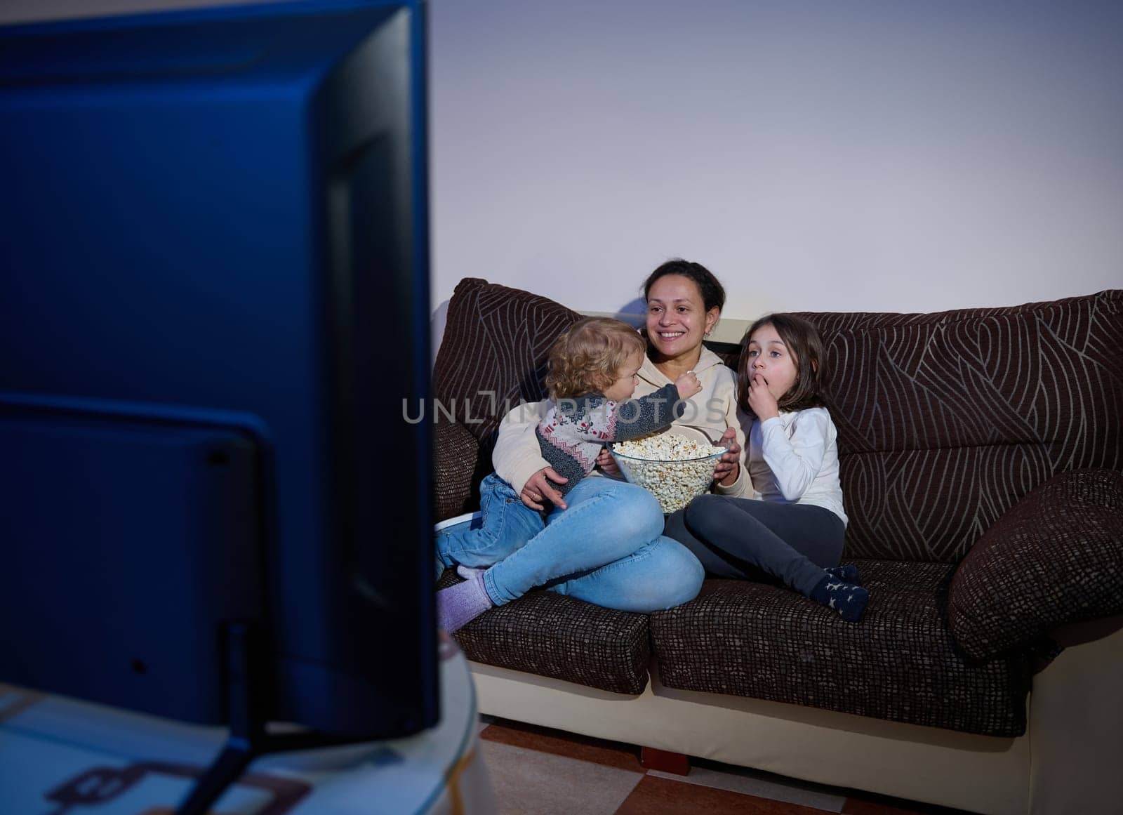 A mother and her children enjoying quality time together while watching TV and eating popcorn. This cozy scene highlights family bonding and relaxation at home during a leisurely evening.