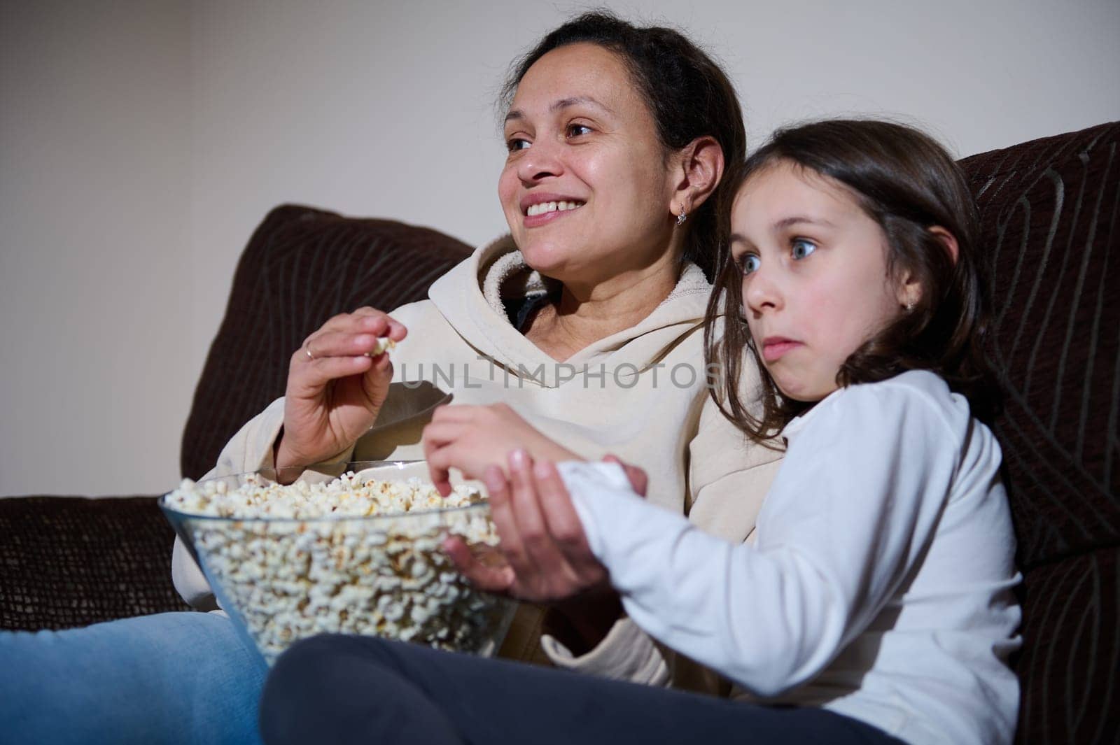 A mother and her daughter sitting on a couch, eating popcorn, and enjoying a movie night together, showcasing family bonding and quality time spent watching a film.