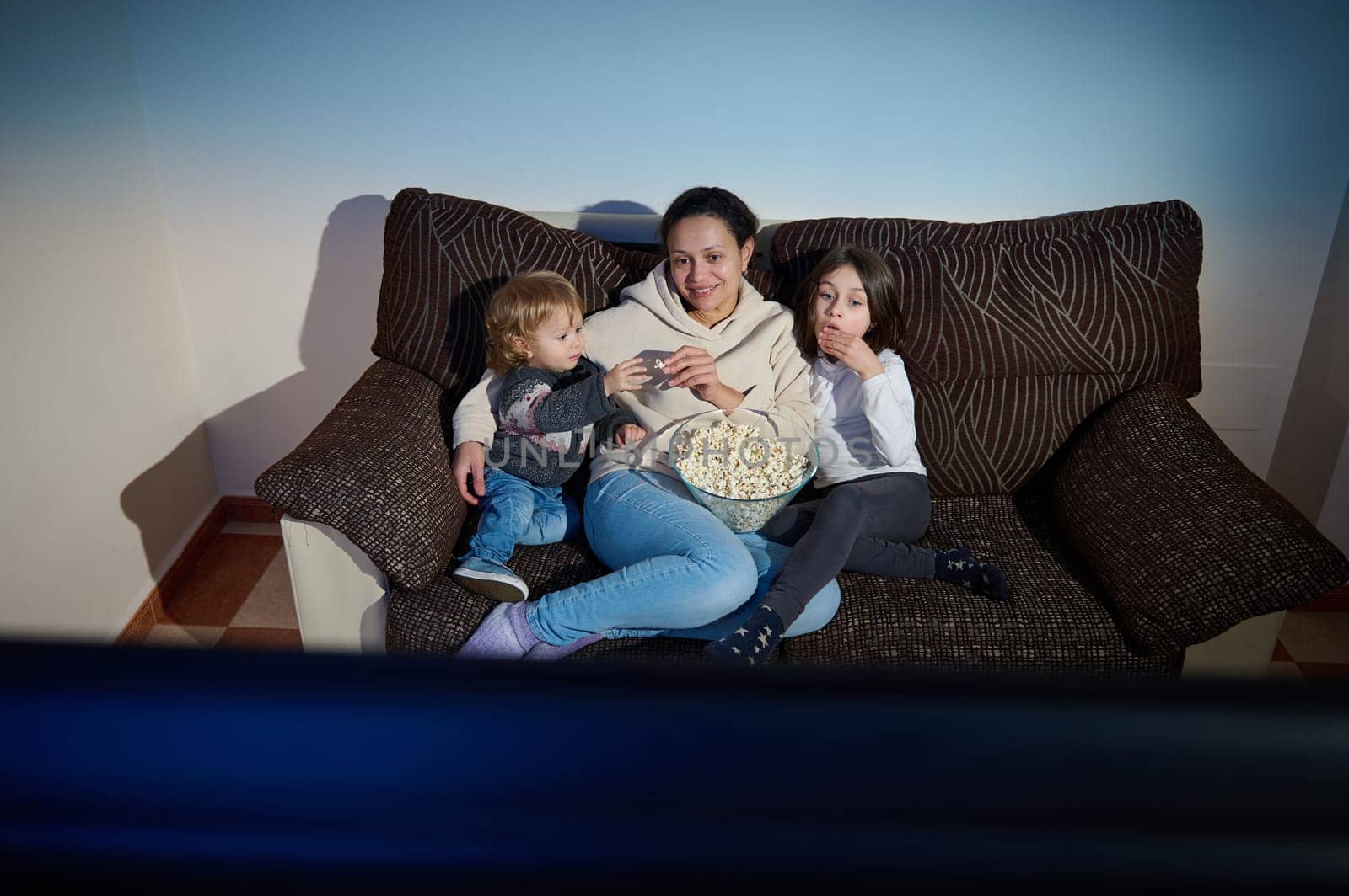 A mother and her two children sit on a couch, sharing popcorn and enjoying entertainment together in a cozy living room setting. Their expressions reflect relaxation, enjoyment, and the warmth of familial bonding.