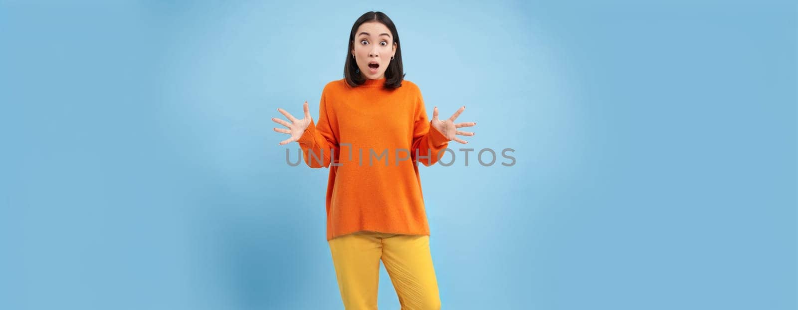 Frustrated young asian woman, shakes hands and looks concerned, panicking, looking worried and upset, standing over blue background.