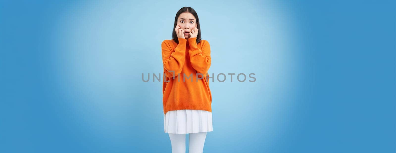 Scared Asian woman shuts her eyes and peeks through hands at smth scary and disturbing, shocked by smth horrifying, standing over blue background.