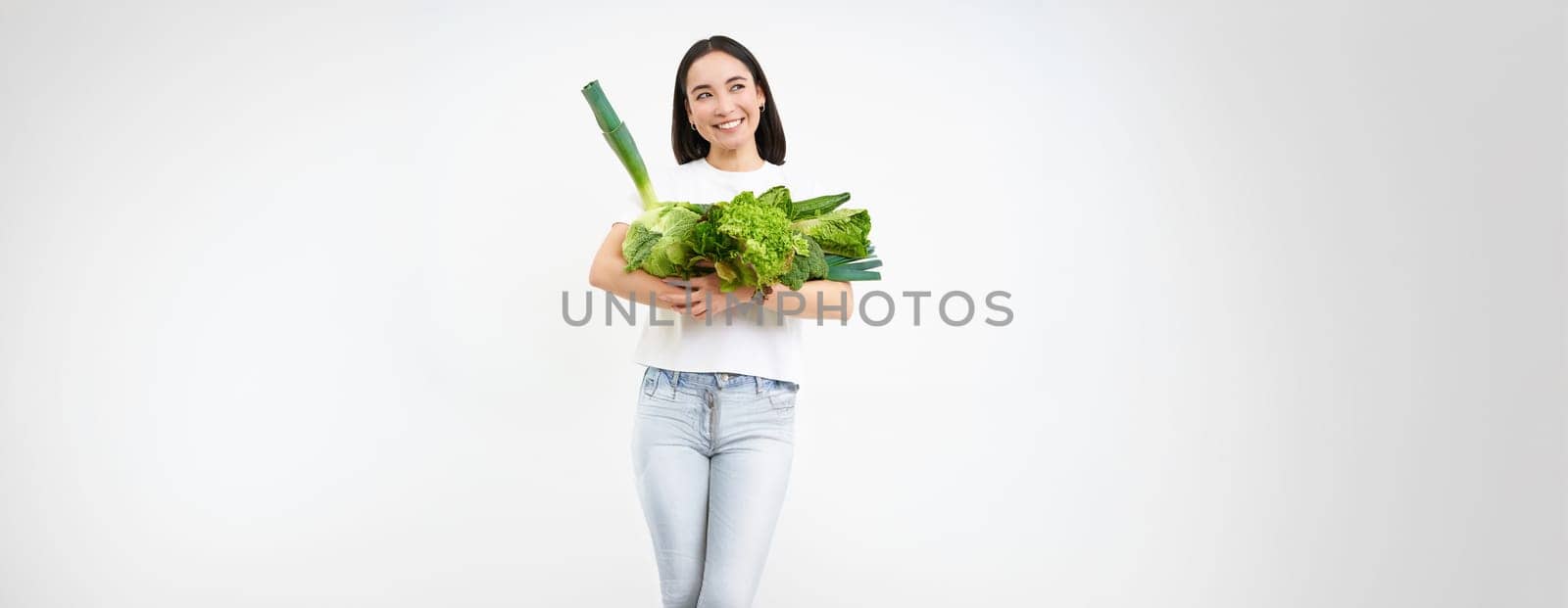 Portrait of happy young woman on green diet, likes eating organic food, holding fresh vegetables from garden, lettuce, cabbage, white studio background.