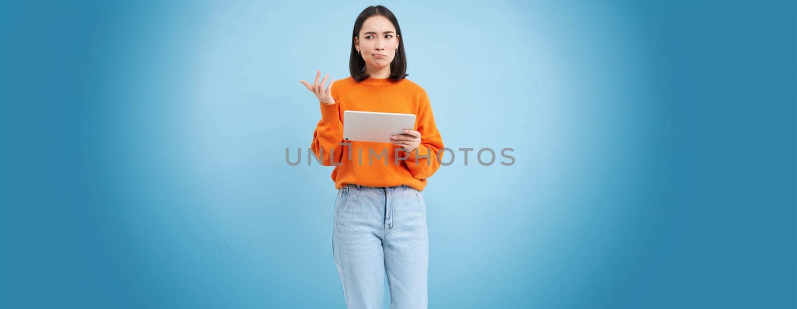 Woman with digital tablet, looks confused, cant understand smth on her gadget, stands puzzled against blue background.