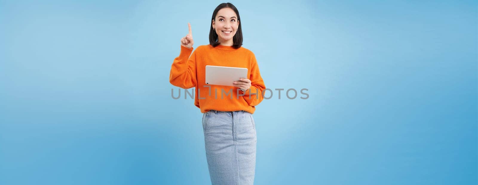 Beautiful young korean woman with tablet, pointing finger up and looking on top advertisement, showing banner, standing over blue background. Online shopping concept.