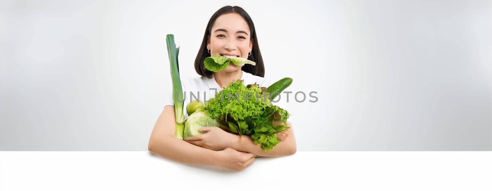 Portrait of smiling korean woman eats lettuce leaf, holding green vegetables, eating nutrisious food, white background.