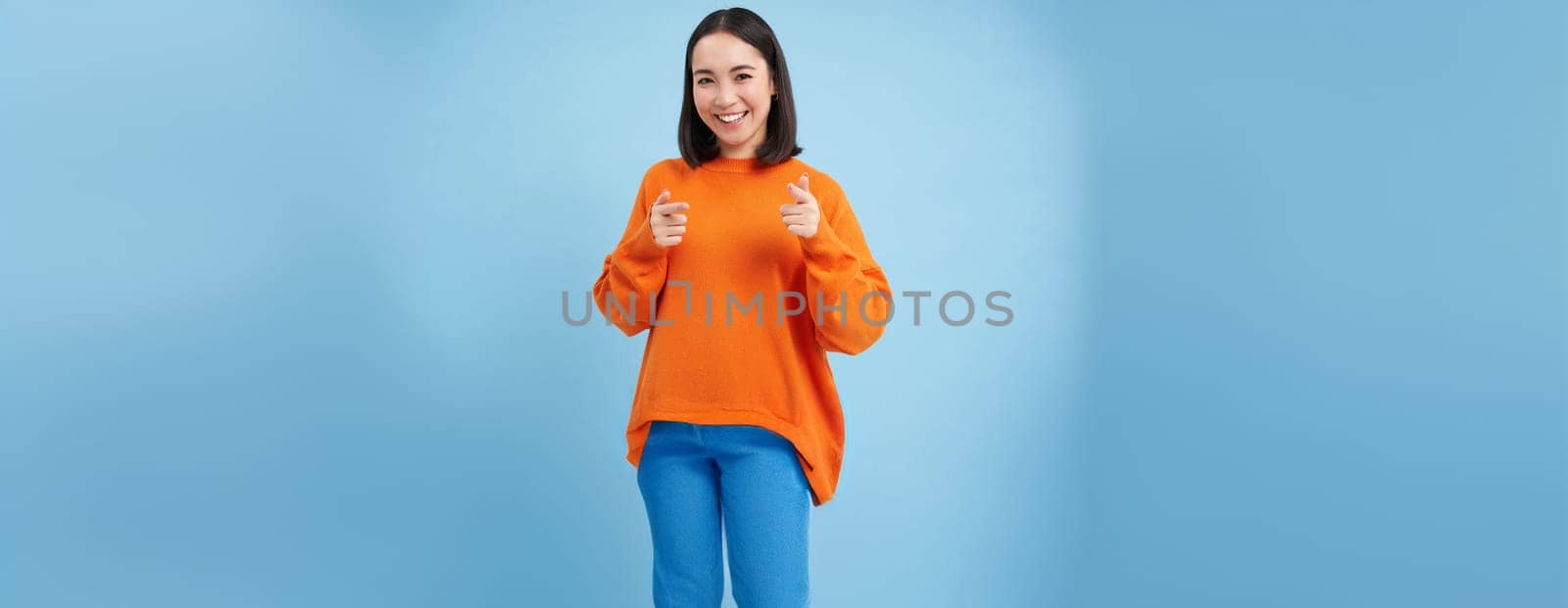 You got this, good job. Enthusiastic korean woman, points at camera with finger pistols and smiles, encourages, praises, stands over blue background.
