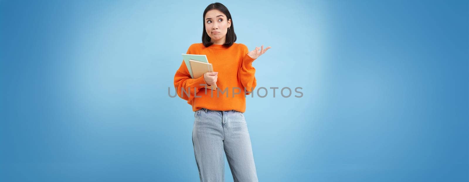 Confused asian woman, student with puzzled face shrugs shoulders, holds notebook, stands over blue background.