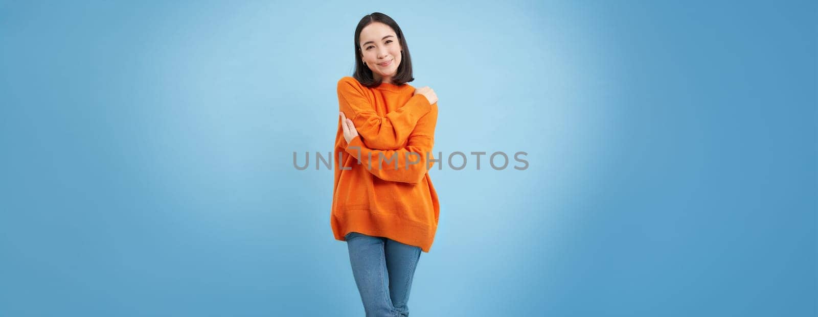 Warmth and tenderness. Cute korean girl hugs herself, embrace equite, femenism and international womens day concept, blue background.
