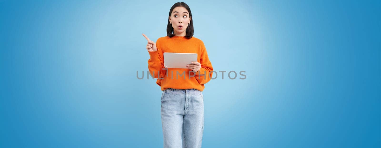 Excited korean woman with digital tablet, pointing at empty space, advertisement banner, holding gadget, showing promo offer, blue background.
