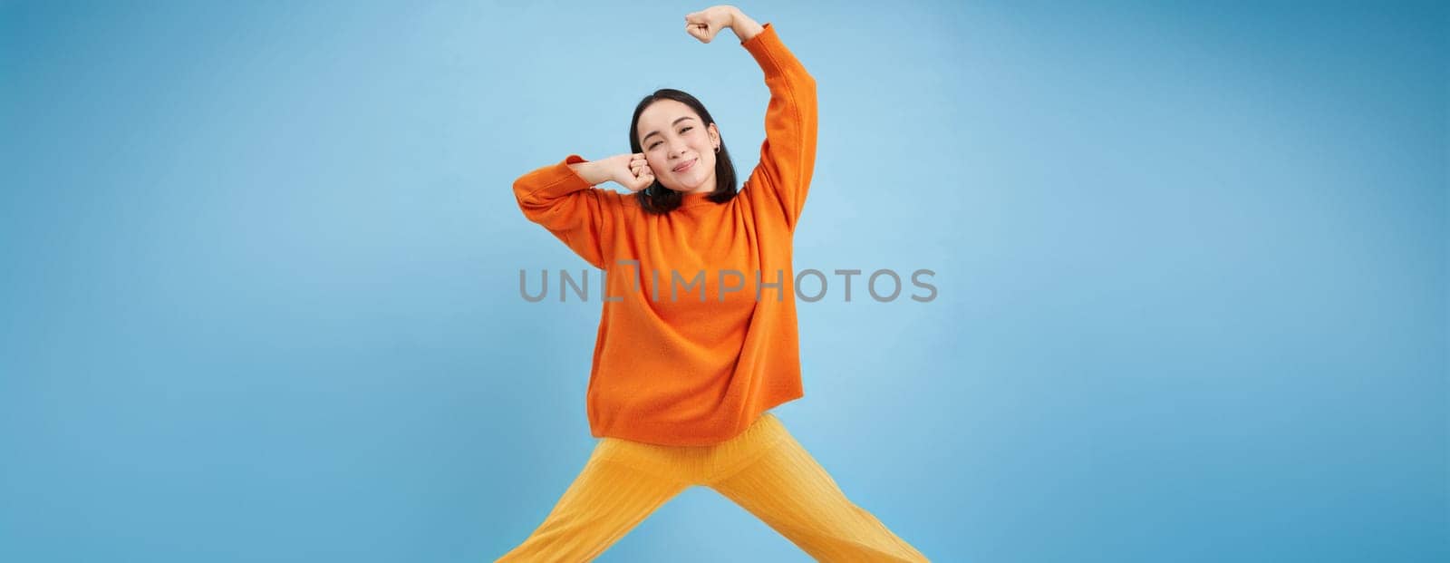 Smiling young korean woman stretching her arms with satisfied face, pleased after good nap, waking up, standing over blue background.