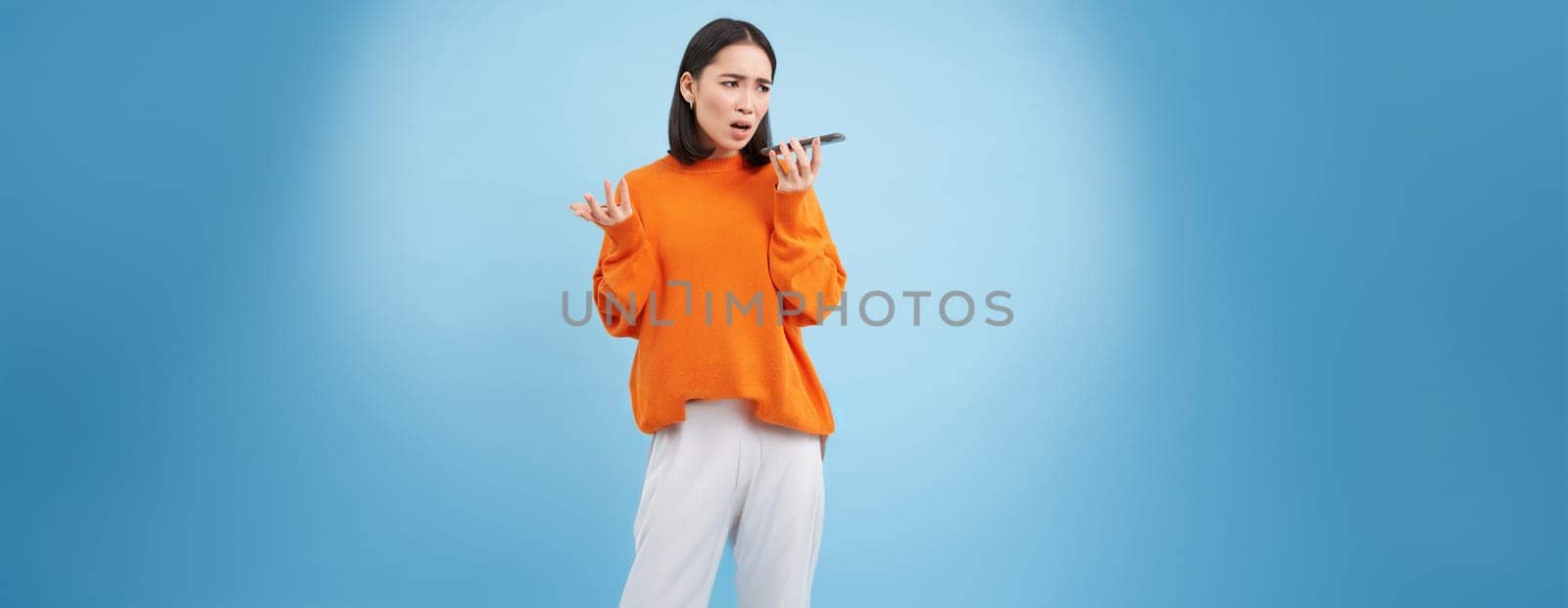 Portrait of upset young woman records voice mail or message, complains with disappointed, sad face, standing over blue background and holding mobile phone.