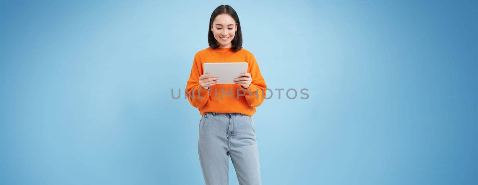 Smiling beautiful asian woman reading on digital tablet, watching videos on gadget or playing video games, standing over blue background.