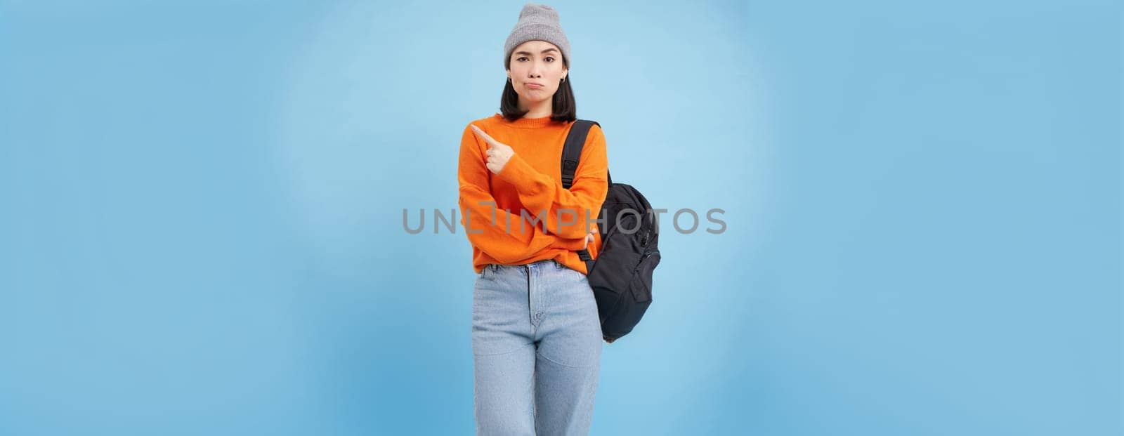 Skeptical asian woman pointing left at strange banner, looks doubtful, stands over studio background.
