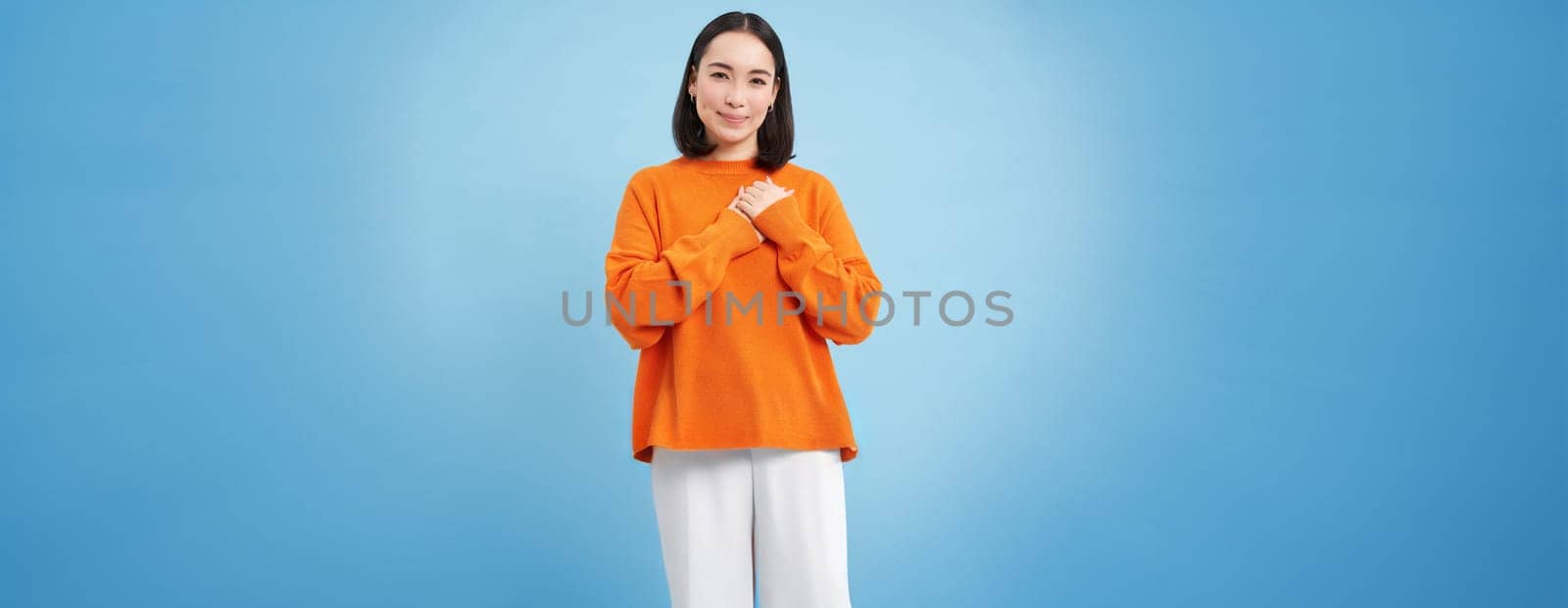 Care and love. Asian woman holds hands on heart and smiles, looks with tenderness and warmth at camera, stands over blue background.