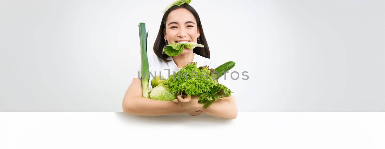 Portrait of smiling korean woman eats lettuce leaf, holding green vegetables, eating nutrisious food, white background.