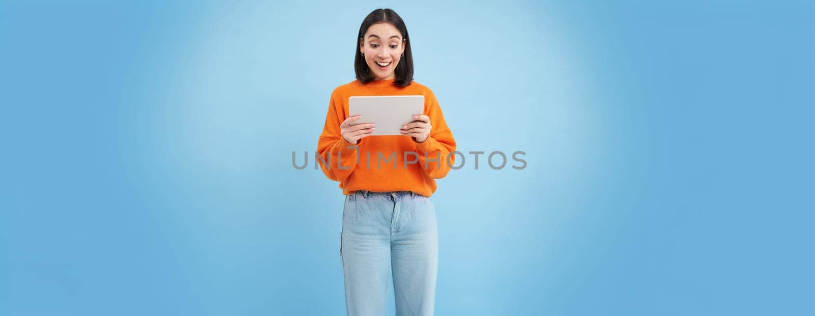 Smiling beautiful asian woman reading on digital tablet, watching videos on gadget or playing video games, standing over blue background.