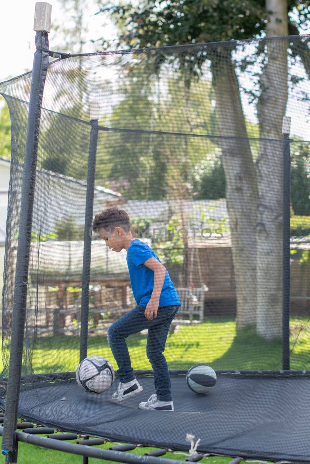 Young boy playing football on trampoline in garden, enjoying the warm summer weather while getting exercise and having fun in the sun, surrounded by greenery and trees,concept of peaceful childhood