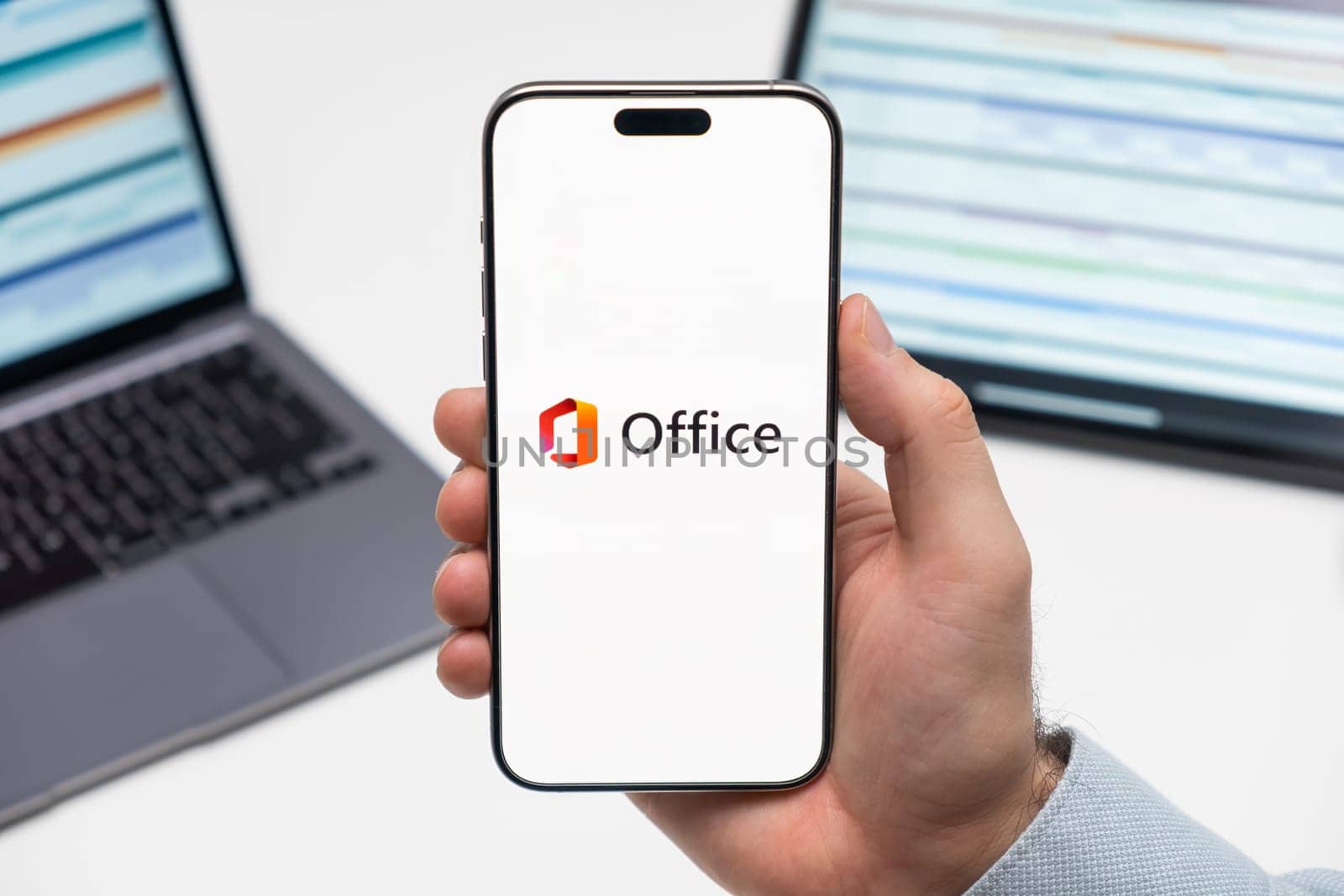 PRAGUE, CZECHIA - JULY, 2024: Microsoft Office logo on the screen of smartphone in mans hand and laptop on the background