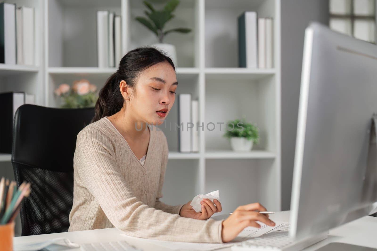A diverse woman in an office, dealing with stress while holding tissues and working on a computer at her desk.