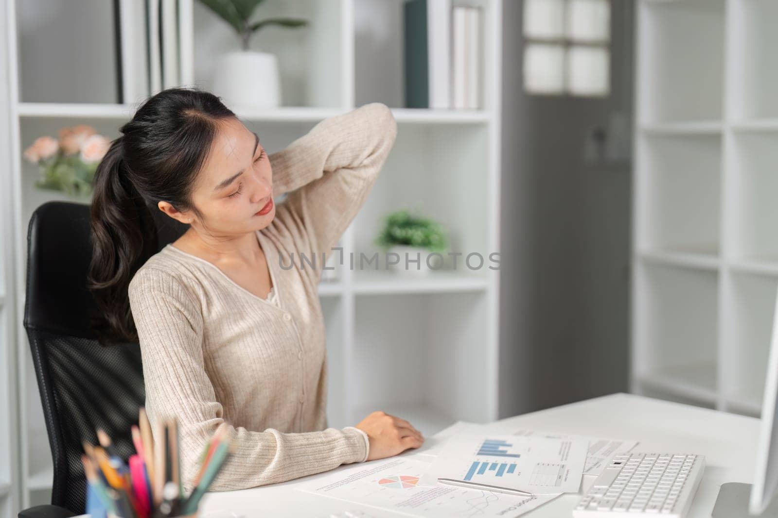 A woman in a modern office experiencing neck discomfort while examining financial reports at her desk.