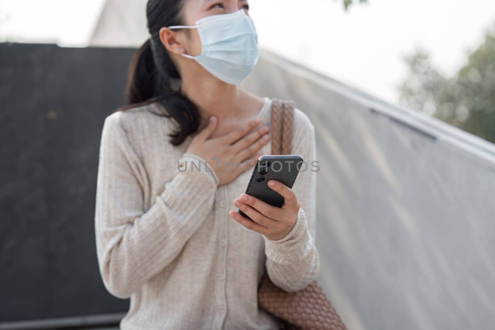 A young woman wearing a protective mask is holding her smartphone close to her chest, expressing emotion while enjoying her time in an outdoor public setting.