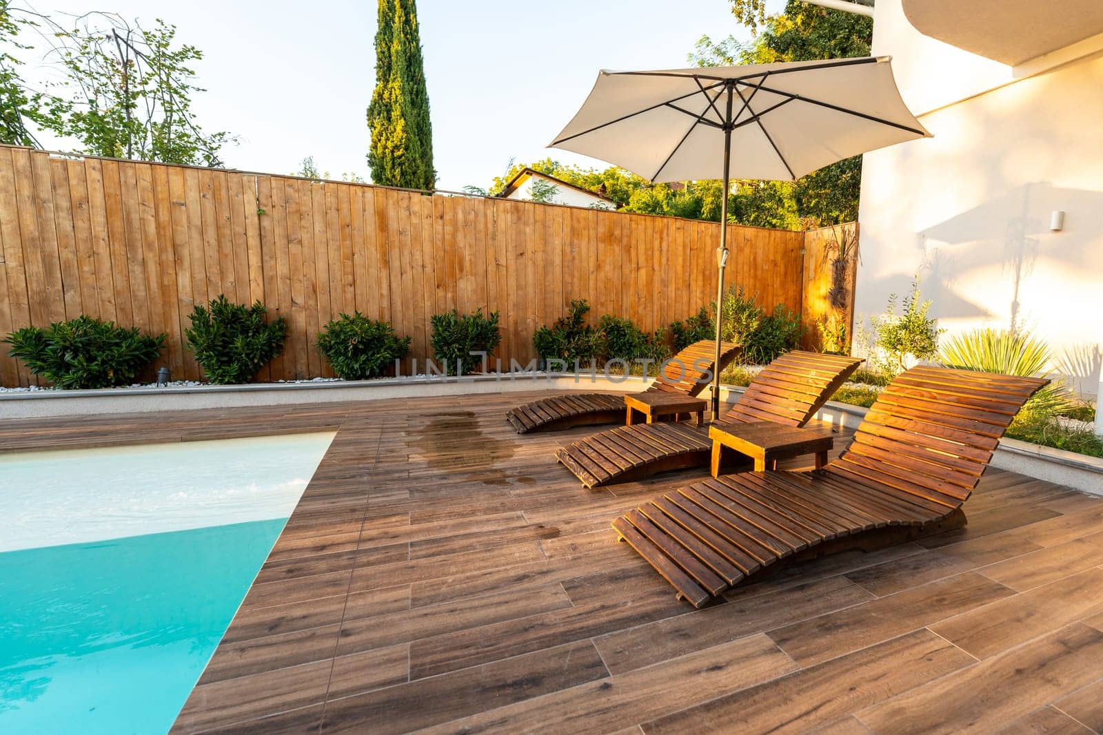 Modern wooden poolside deck with curved lounge chairs, umbrella, and turquoise pool in a sunny backyard