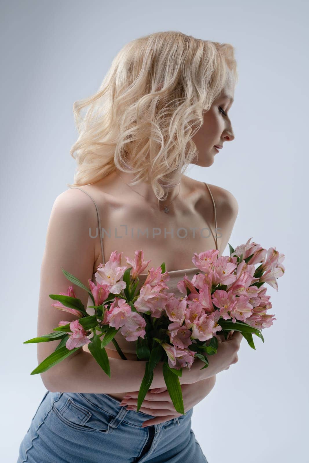 A fair-haired model stands elegantly, holding a bouquet of pink flowers, creating a soft and serene atmosphere in the studio.