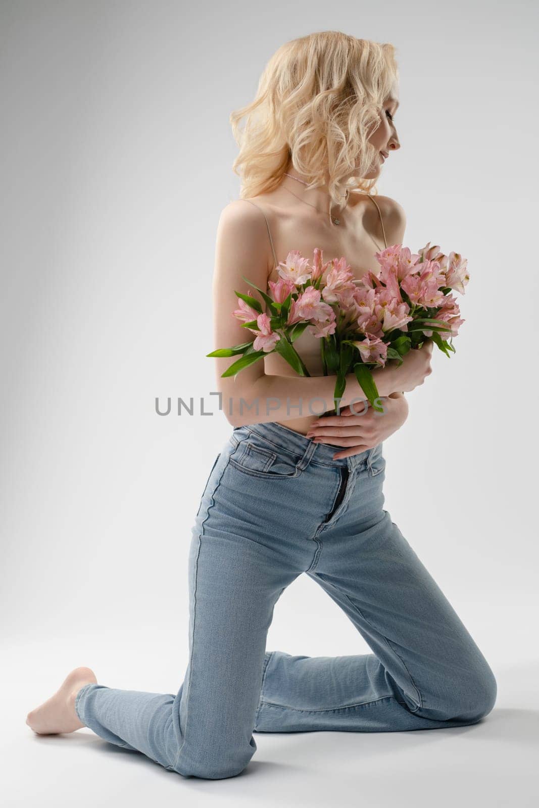 A woman kneels with elegance, holding a bouquet of pink flowers against a neutral backdrop, highlighting her natural beauty and style.
