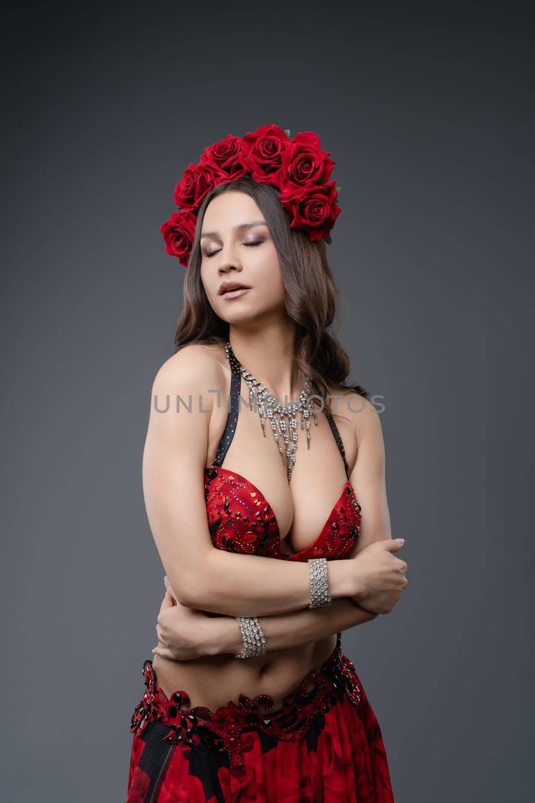 A dancer adorned in red attire and a crown of flowers poses with elegance, showcasing her expressive movements against a neutral background.