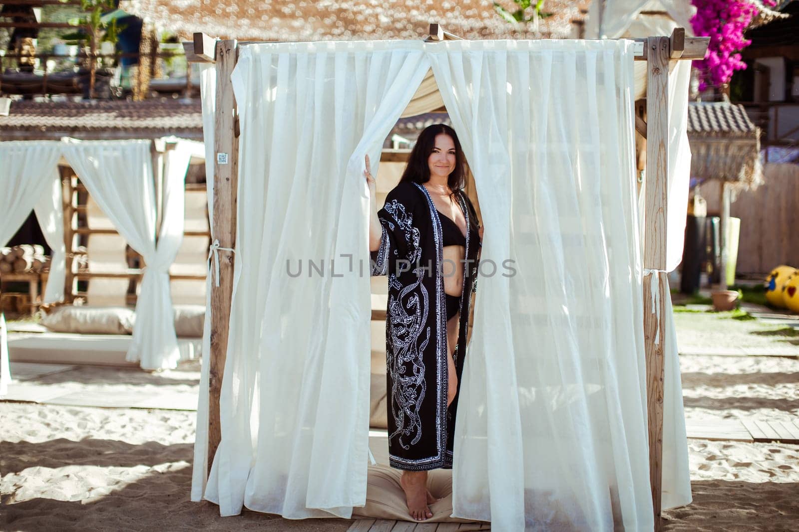 A brunette girl in a black cape with an oriental ornament stands in a beach tent