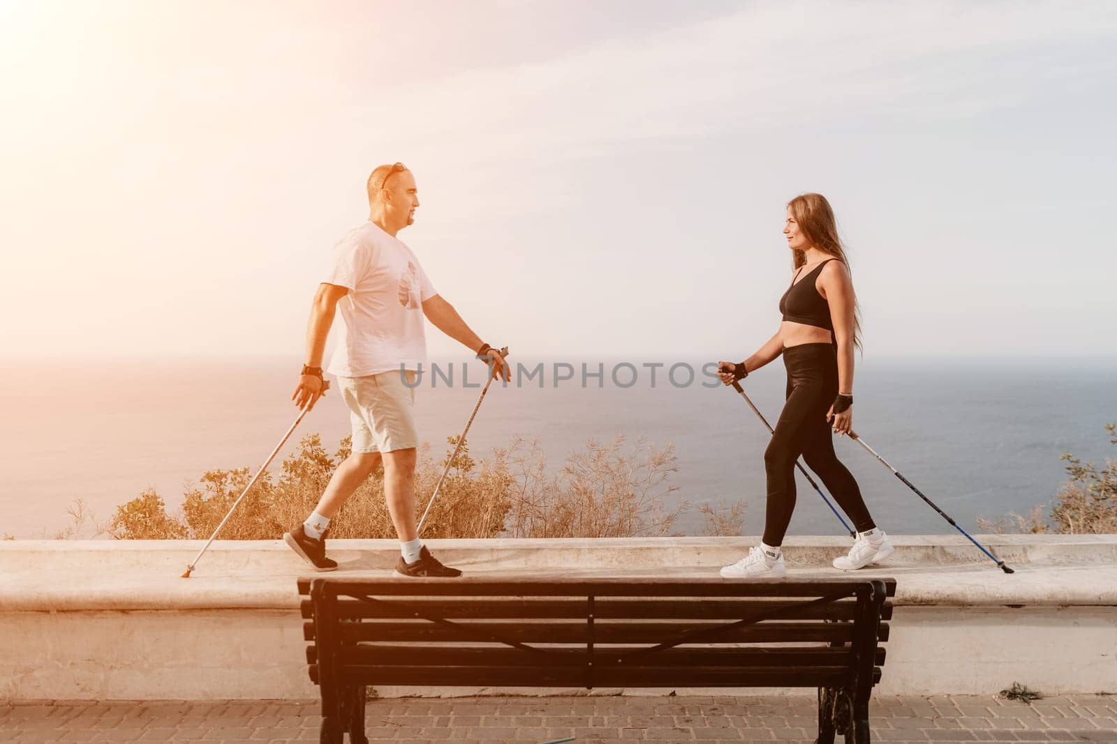 Happy Middle aged couple or friends practicing nordic walking in park near sea. Mature couple with trekking poles walking, practicing Nordic walking outdoors. Aging youthfully and sport concept by panophotograph