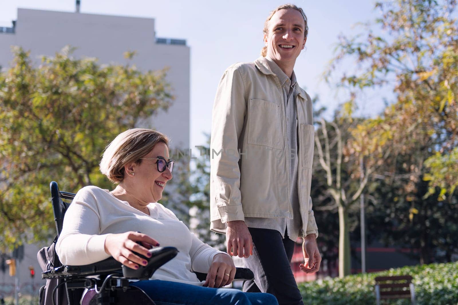portrait of a woman using electric wheelchair with a young man talking happy as they walk through the city, concept of friendship and diversity