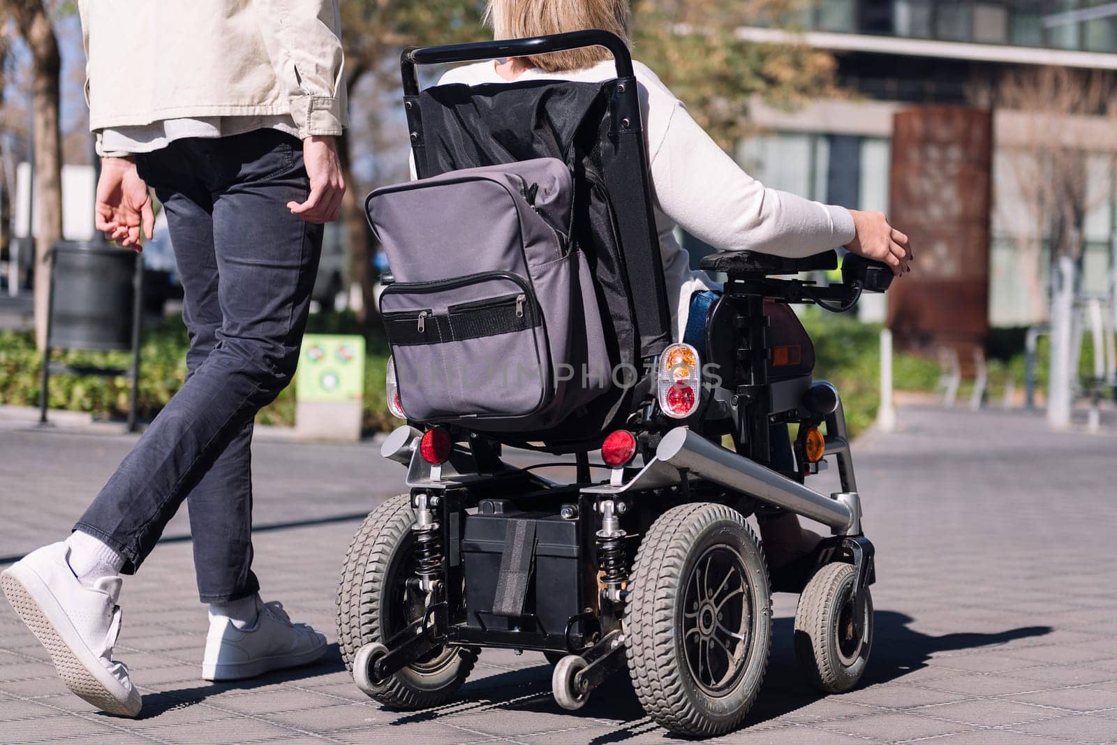 cropped rear view of an unrecognizable woman using wheelchair with a man taking a walk by the city, concept of friendship and diversity