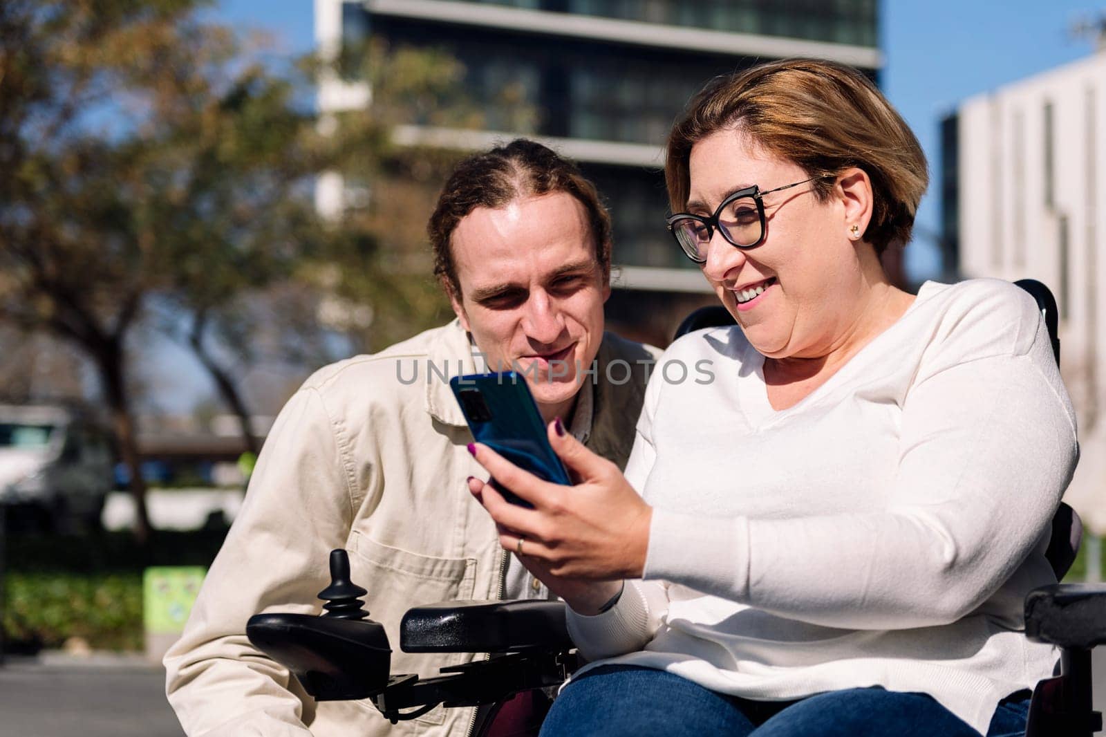 smiling woman using wheelchair showing mobile phone to a young male friend, concept of friendship and technology of communication