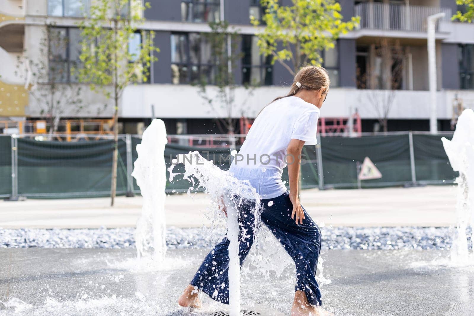 Cheerful young teen girl in city fountain, girl in wet clothes is having fun and enjoying the cool summer water, background city architecture. High quality photo
