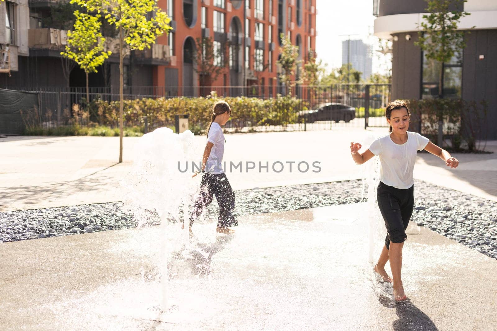 Cheerful young teen girl in city fountain, girl in wet clothes is having fun and enjoying the cool summer water, background city architecture. High quality photo