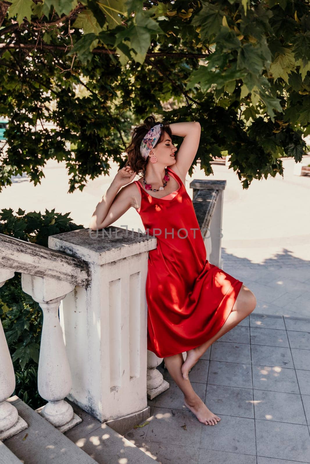 woman in a red silk dress and a bandage on her head smiles against the background of the leaves of a tree. She is leaning on the coop and looking into the camera. Vertical photo