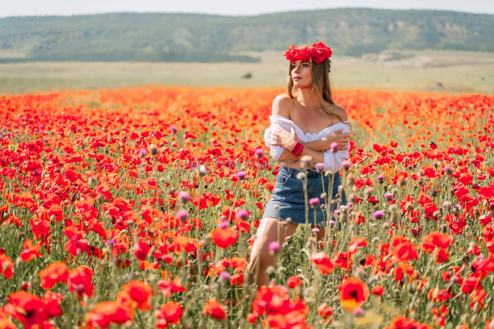 Happy woman in a poppy field in a white shirt and denim skirt with a wreath of poppies on her head posing and enjoying the poppy field. by Matiunina