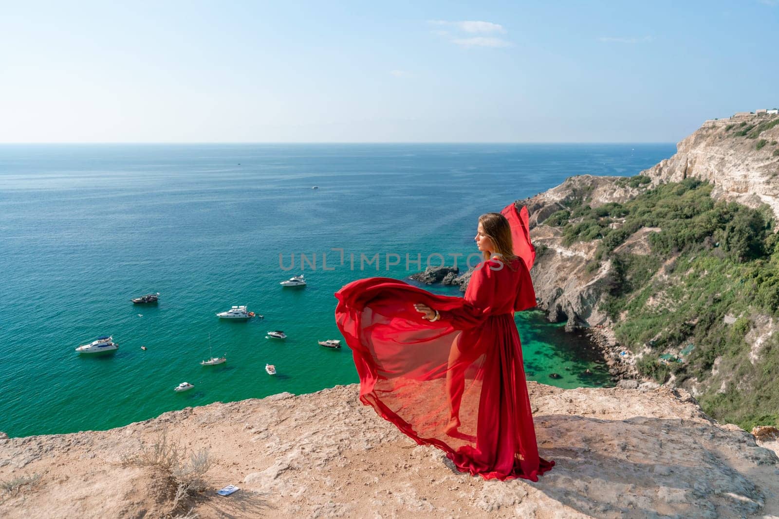 Red Dress Woman sea Cliff. A beautiful woman in a red dress and white swimsuit poses on a cliff overlooking the sea on a sunny day. Boats and yachts dot the background