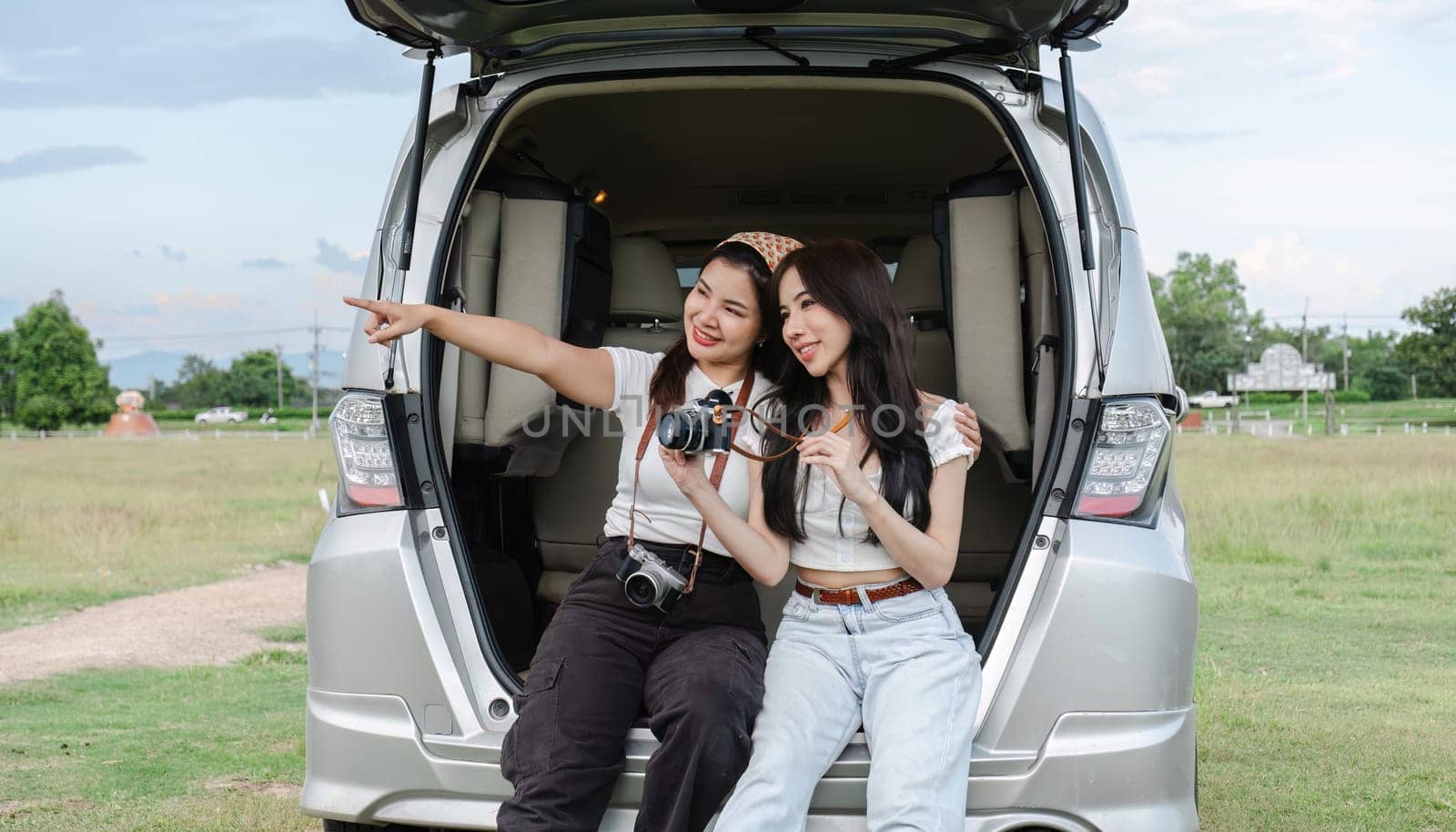 Two young Asian women in white shirts and jeans sit in the back of their new car, lounging on the field. holidays and traveling by wichayada