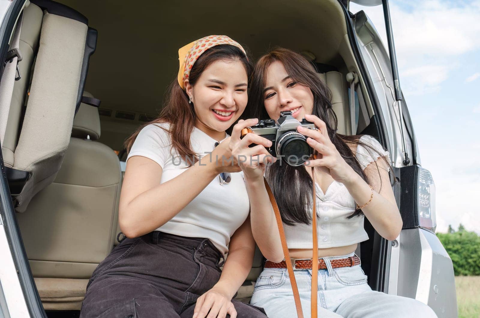 Cute Asian female friends enjoying taking photo with a camera together during a trip. and enjoy sitting and chatting in the back of the car on holiday.