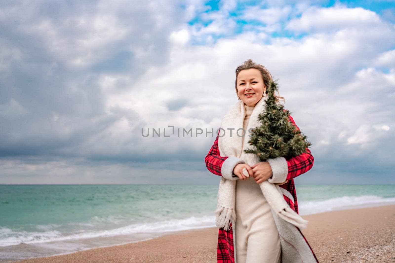 Blond woman holding Christmas tree by the sea. Christmas portrait of a happy woman walking along the beach and holding a Christmas tree in her hands. Dressed in a red coat, white dress. by Matiunina