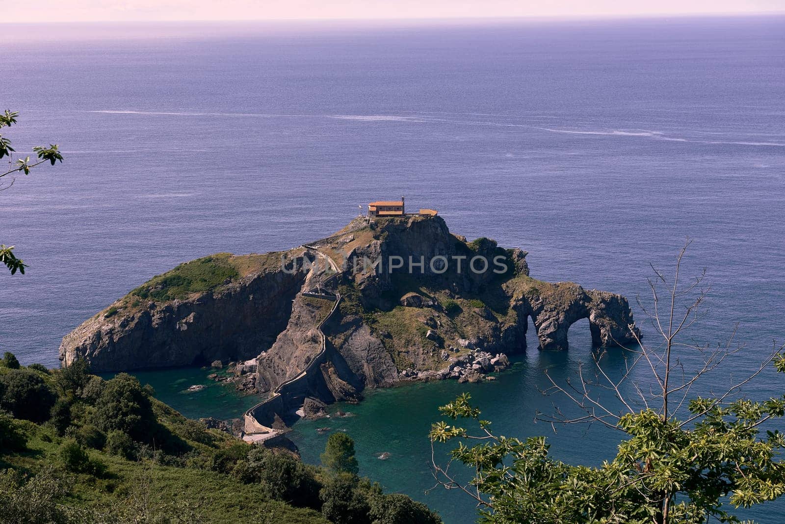 Hermitage of San Juan de Gaztelugatxe on a sunny day. calm sea, rocky formation, holes, frame of branches and trees