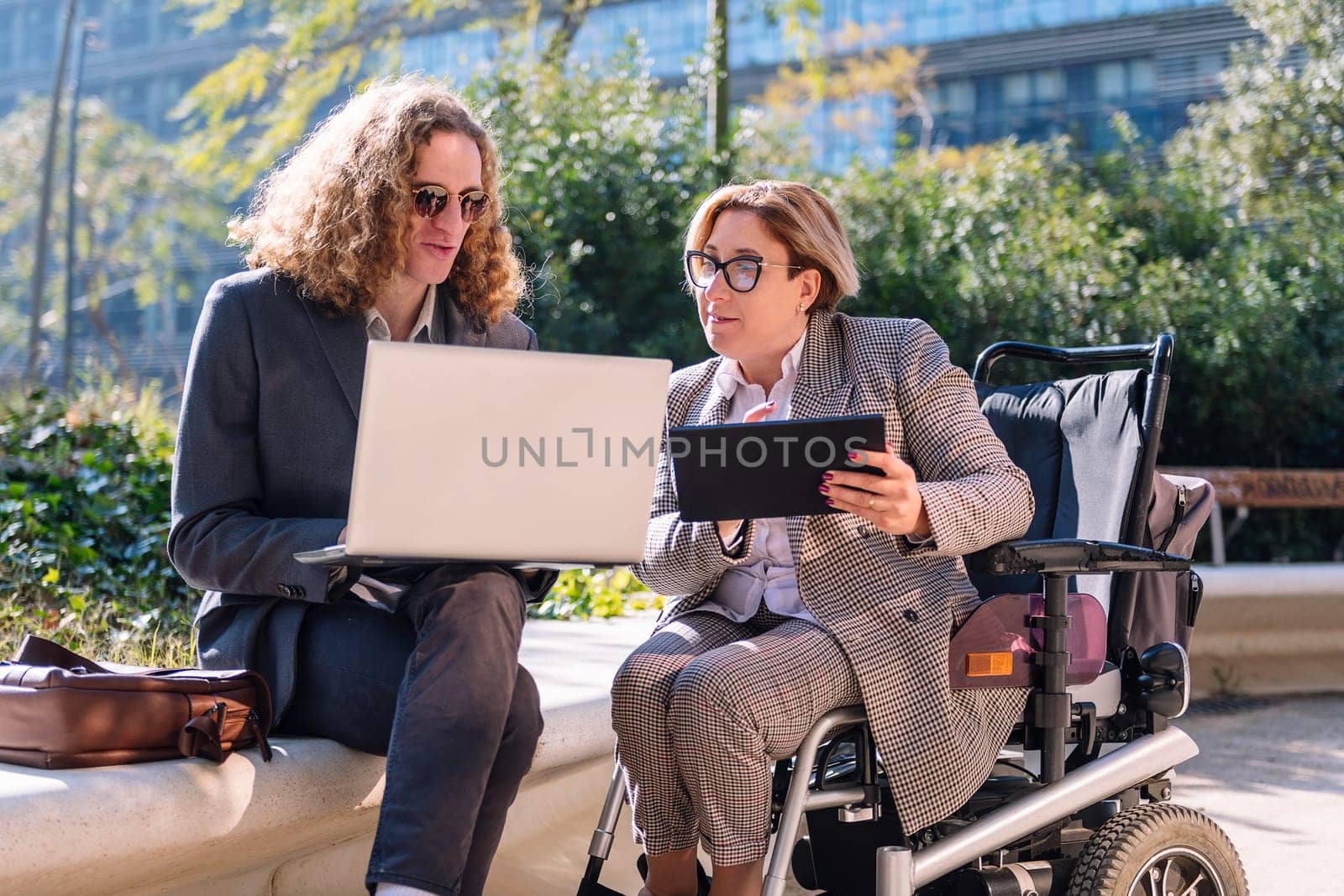 business man and woman using wheelchair working outdoors with laptop and tablet, concept of diversity and urban lifestyle